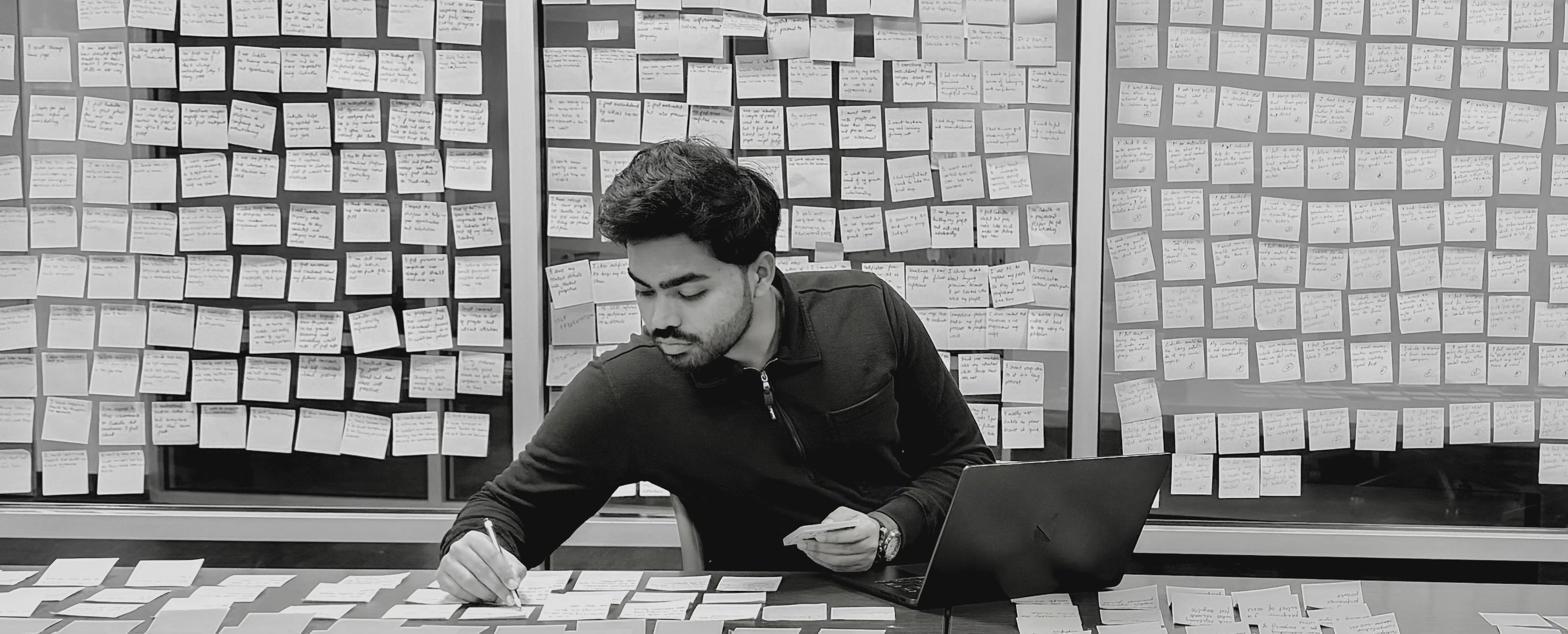 Arjun writing at a table in front of a wall of research notes